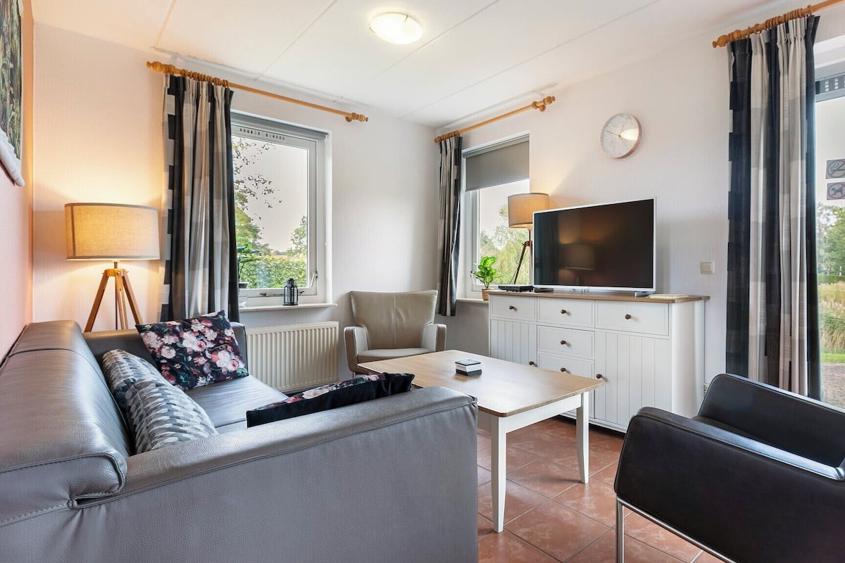 Een moderne woonkamer met een bank, salontafel, televisie en een stoel. Er is een raam met gordijnen, een radiator en een klok aan de muur.