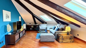 Ein gemütlicher Dachbodenwohnbereich mit hohen Decken, Holzbalken, einem Fernseher, einem Sofa, einem Couchtisch und einem Koffer.