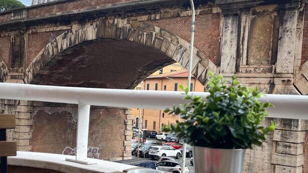 A balcony with a wooden table, two red cups, and a potted plant. A bridge and a dome structure are visible in the background. - Ele rooms San Pietro (Rome)