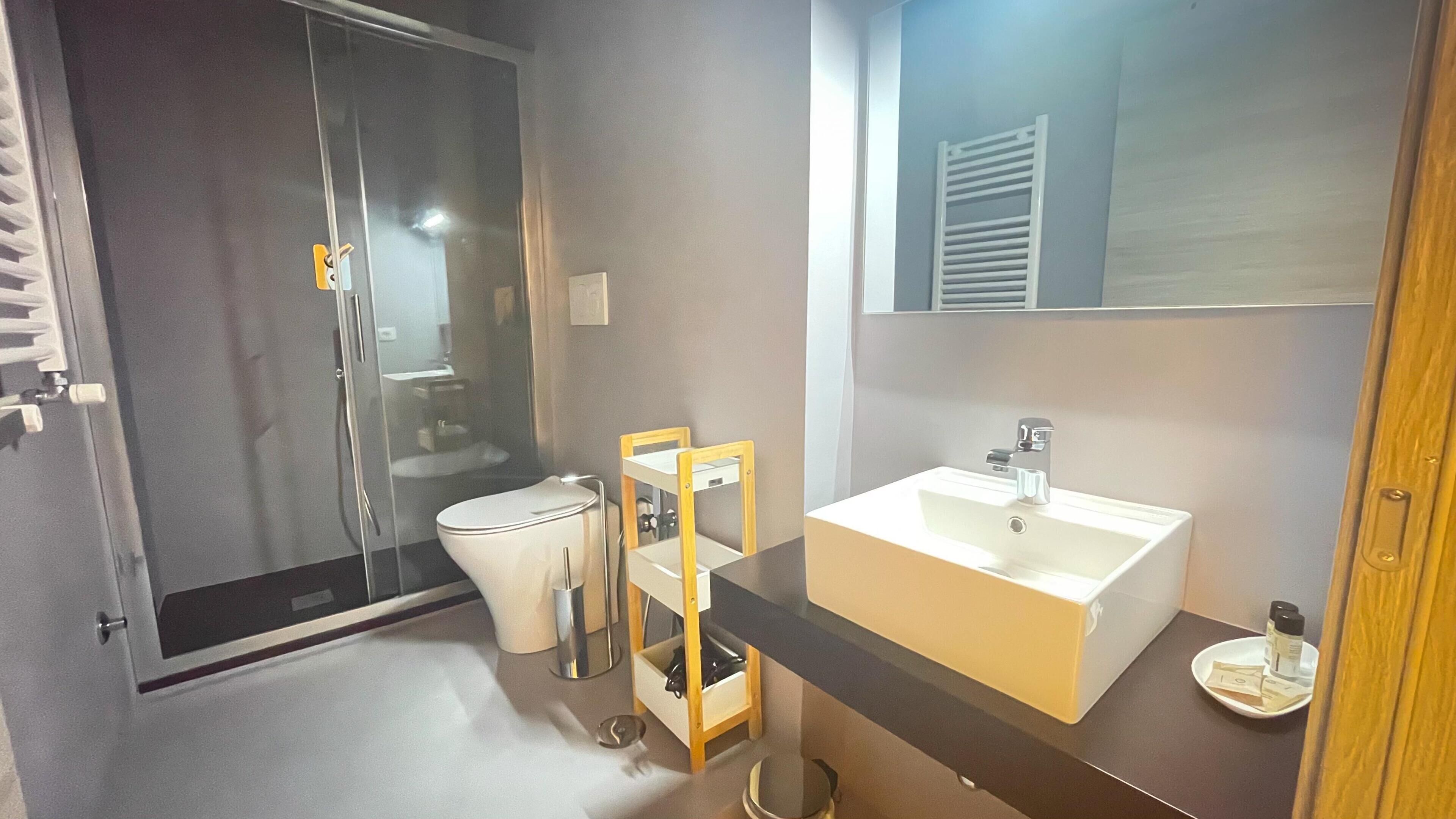A modern bathroom with a glass-enclosed shower, a white toilet, a wooden chair, a white sink, and a mirror above the sink.