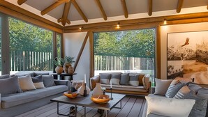 Amplia zona de patio con techo de madera, ventanas grandes y un espacio de descanso que incluye un sofá y una mesa con un cuenco y velas.