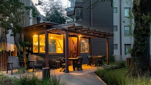Un espacio al aire libre acogedor con mesas y sillas, rodeado de follaje y un edificio al fondo.