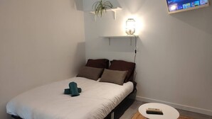 A neatly made bed with a white duvet and brown pillows, a small round table with a remote control, and a wall-mounted TV displaying various channels.