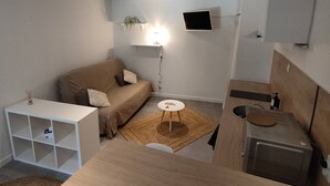 A compact living space with a sofa, a small round table, a kitchenette, and a mounted TV.