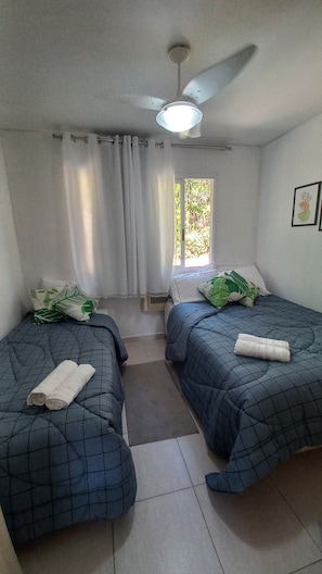 Una habitación con dos camas, ventilador de techo, ventana con cortinas y un pequeño cuadro en la pared.