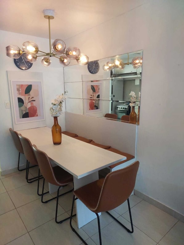 A dining area with a long table, four chairs, a vase with flowers, a clock, and a mirror.
