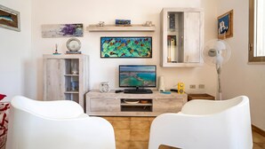 A living room with a TV, white chairs, a cabinet, and artwork on the walls. - Dimora Del Mare - Villa With Garden Near The Sea (Porto Cesareo)