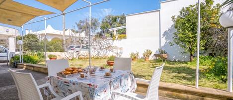 Un espace de restauration en plein air avec une table dressée pour quatre personnes, entourée de chaises en osier blanc, sous une grande tonnelle.