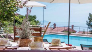 Une table dressée avec une plante, un pot et deux tasses, donnant sur une piscine avec des parasols et offrant une vue sur des bâtiments.