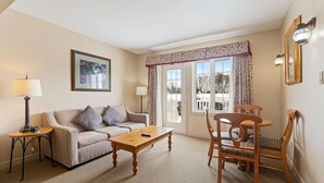 A living room with a sofa, coffee table, and dining table with chairs. There is a window with curtains, a lamp, and a framed picture on the wall. - Peaceful Escape King Bed and Outdoor Activities (Shanty Bay)