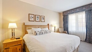 A hotel room with a large bed, two bedside tables with lamps, a wooden nightstand, a chair, and a window with curtains. - Resort Style w/ Balcony, Hot Tub and Full Kitchen (Shanty Bay)