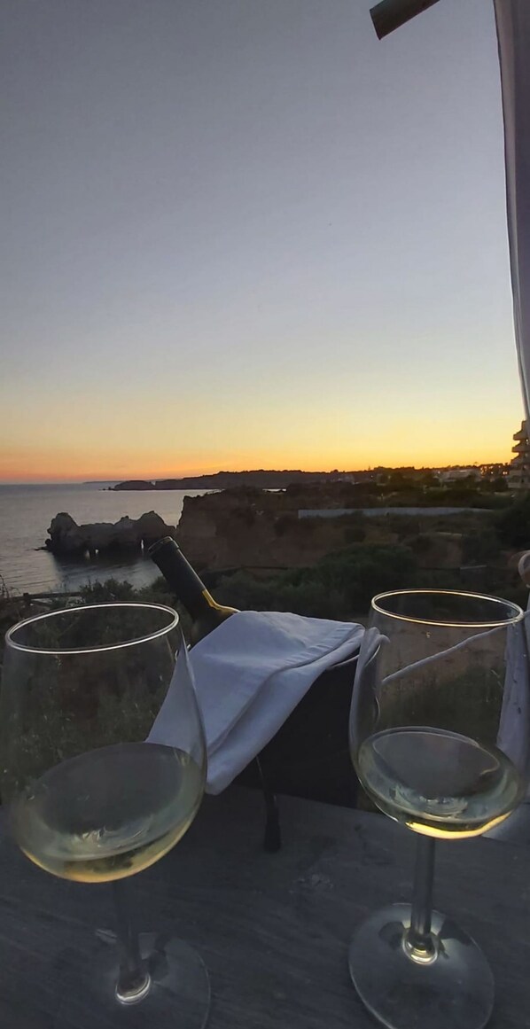 Två vinglas står på ett bord med utsikt över havet och klipporna i solnedgången.