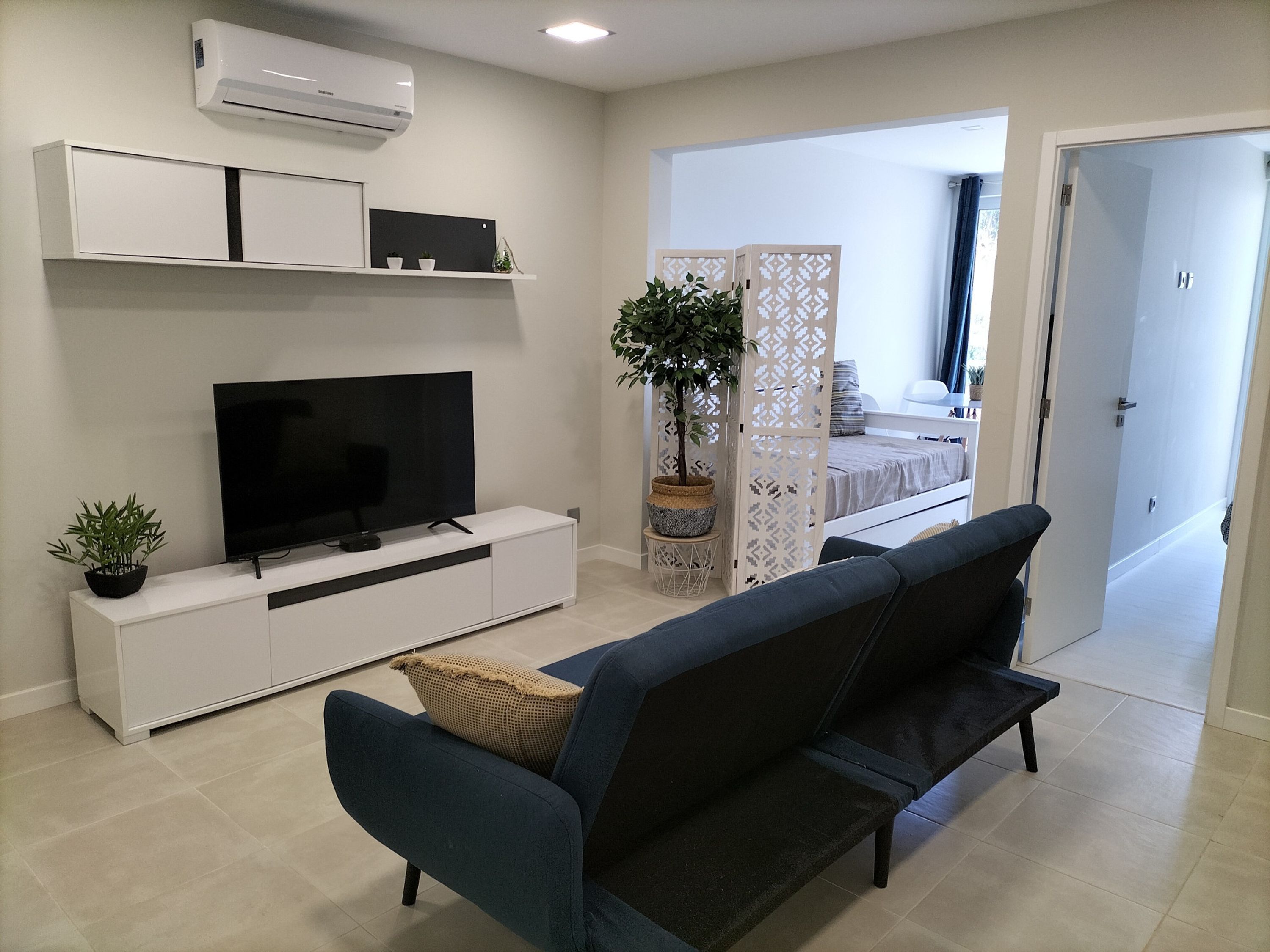 A modern living room with a flat-screen TV on a white console, a blue sofa, and a potted plant.