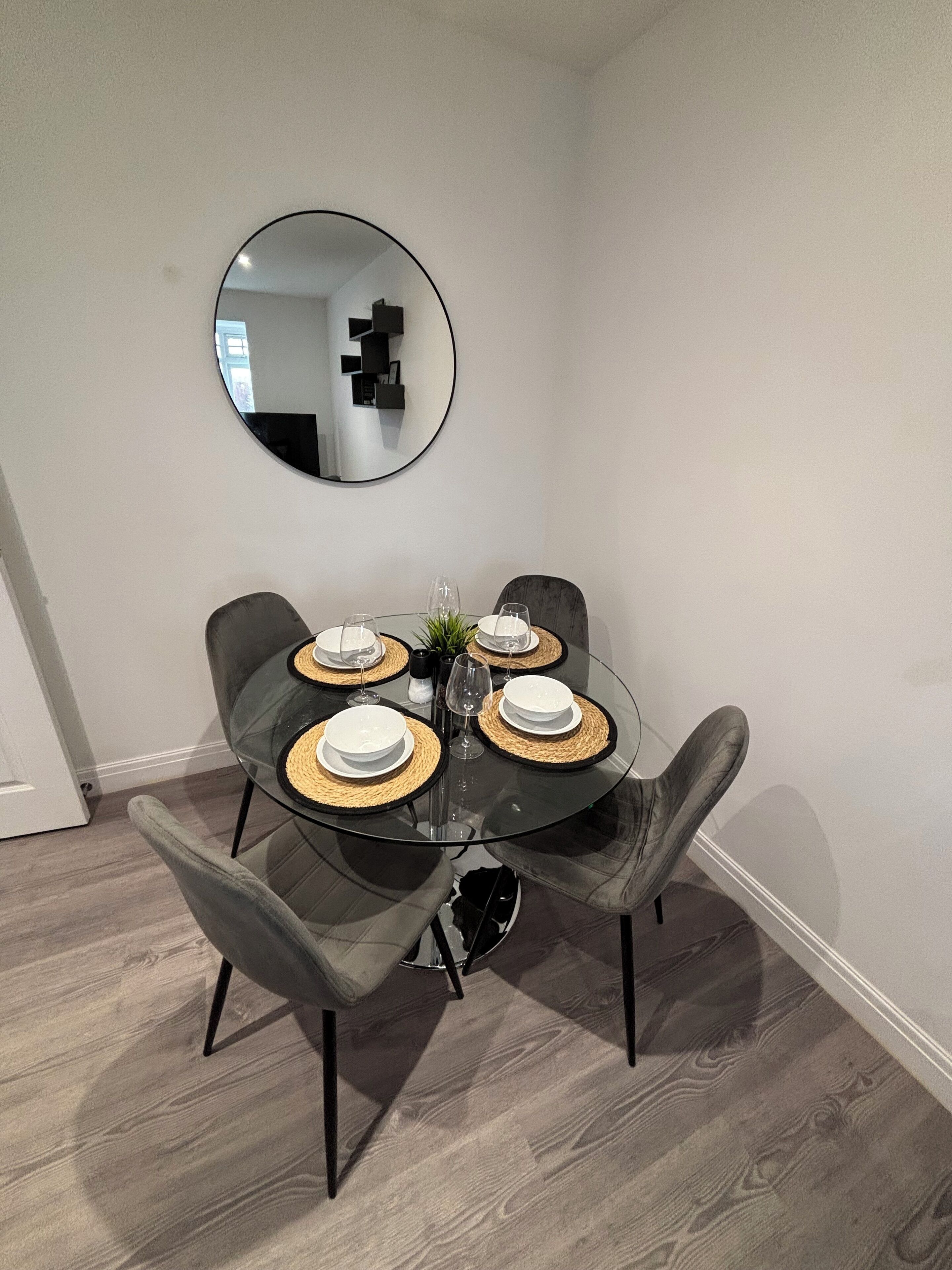 A dining area with a round glass table, four grey upholstered chairs, and a round mirror on the wall.