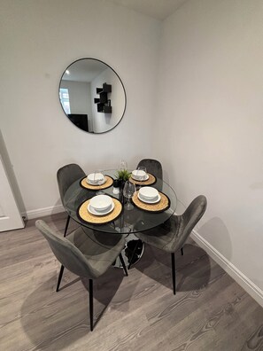 A dining area with a round glass table, four grey upholstered chairs, and a round mirror on the wall.