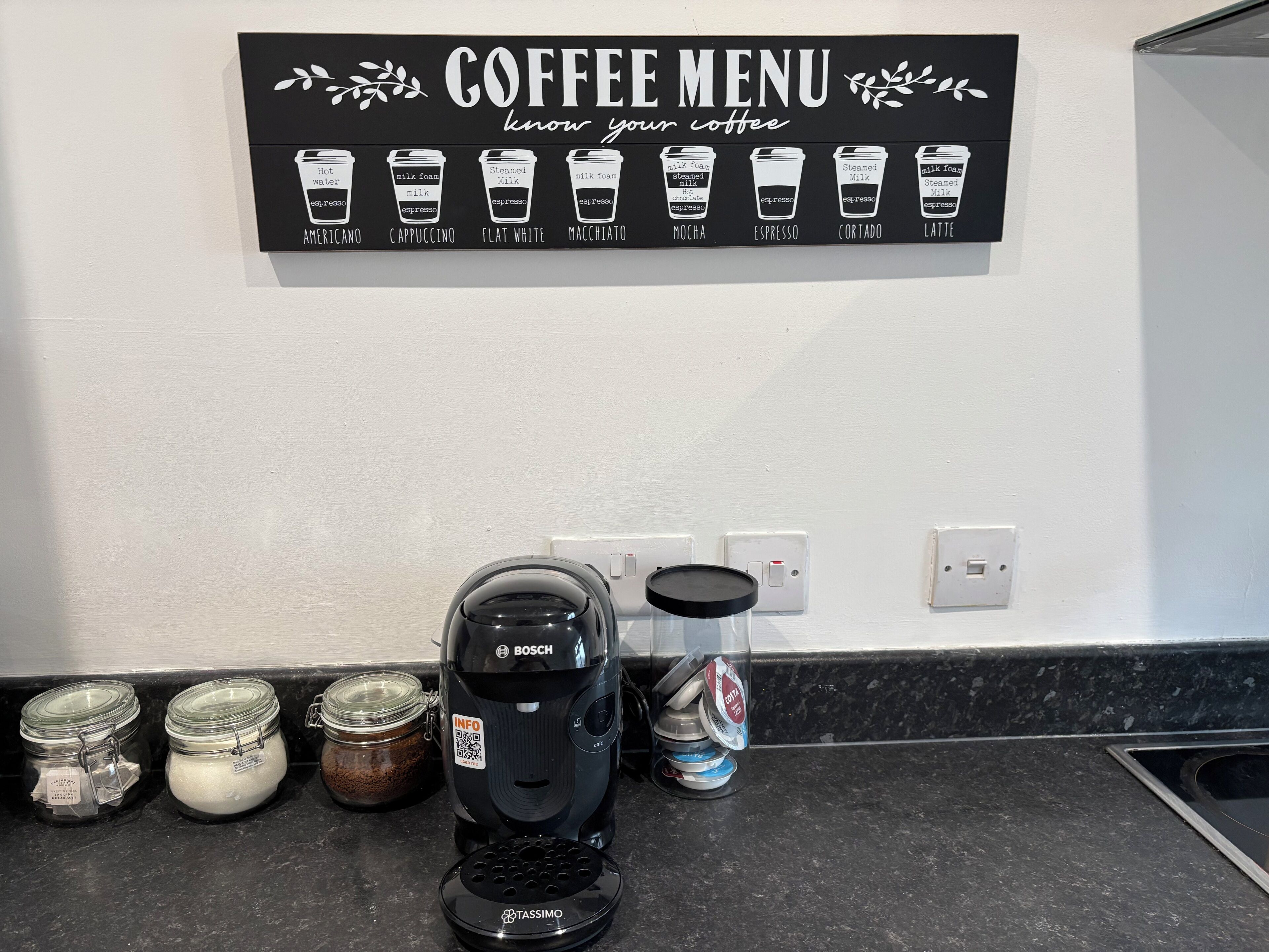 A coffee menu on a wall, a coffee maker, and jars of coffee and cream on a counter.