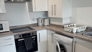 A modern kitchen with white cabinets, a stainless steel range hood, a built-in oven, a washing machine, and a coffee maker.