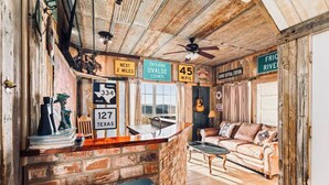 Een rustiek interieur met een bar, een bank en een plafondventilator. Borden geven aan: 'Over 3 kilometer', 'Betreding van Uvalde County', en '64 km/u'