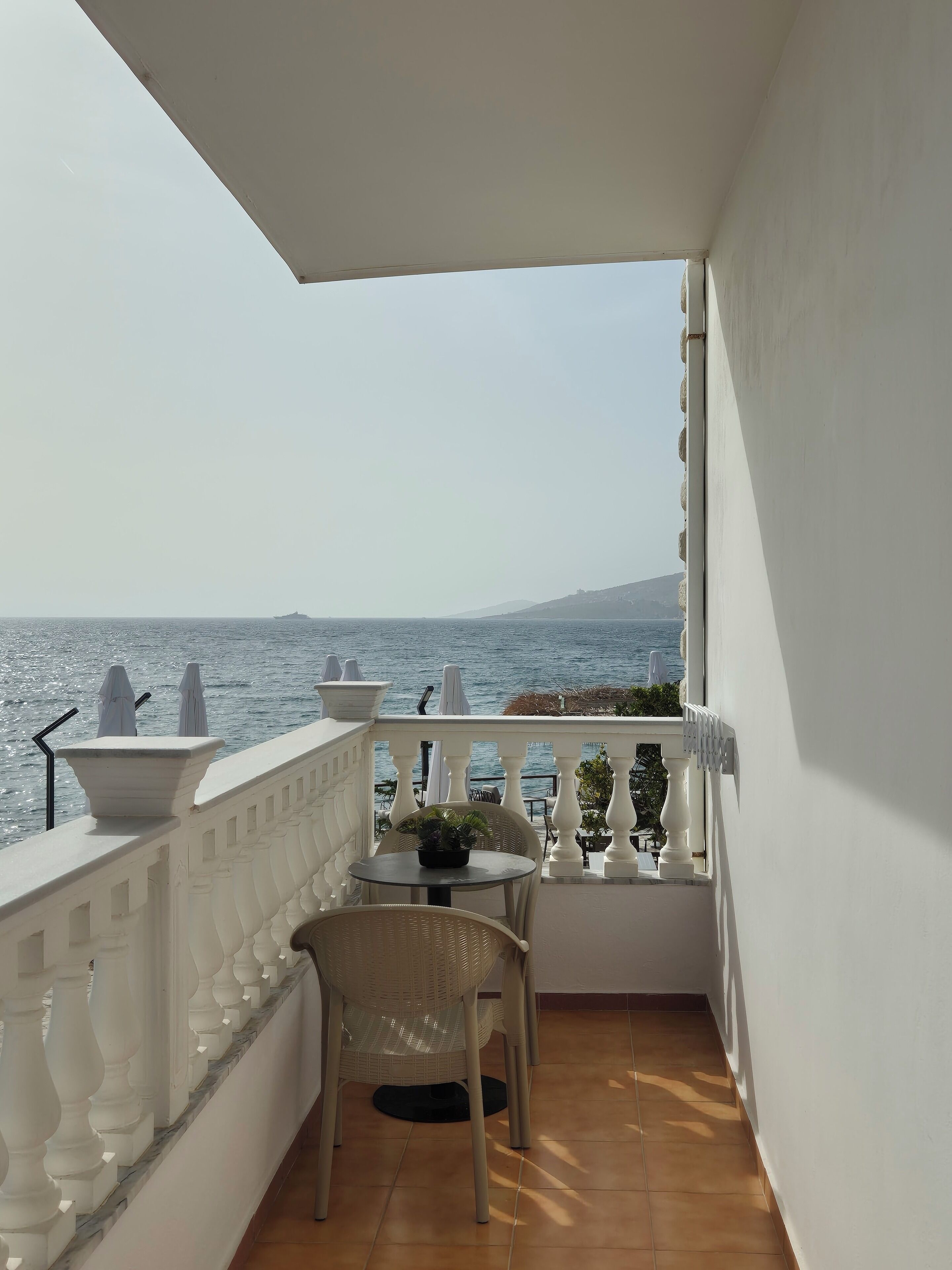 A balcony with a view of the sea, white balustrades, a table with a potted plant, and wicker chairs.