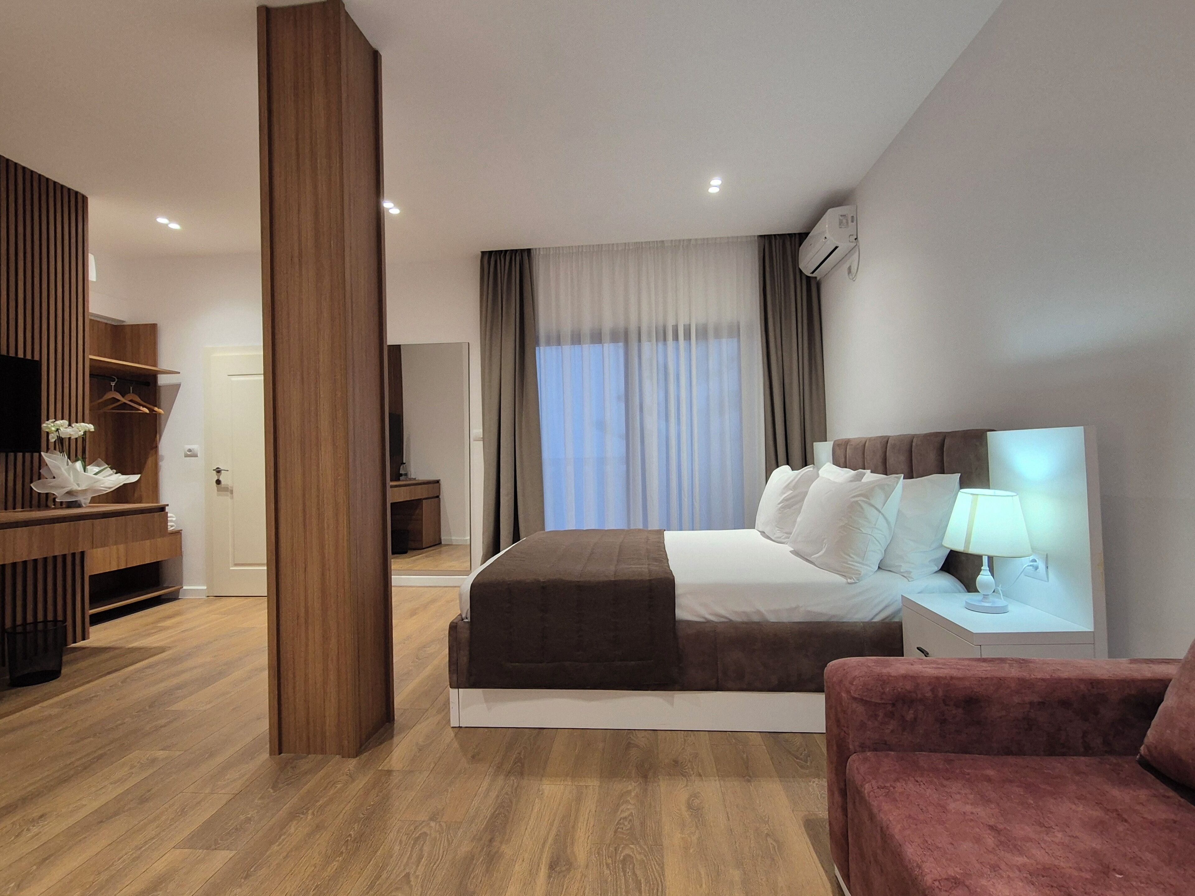 Una habitación de hotel moderna con una cama grande, una mesita de noche con lámpara, un sillón rojo, un armario de madera y un espejo.