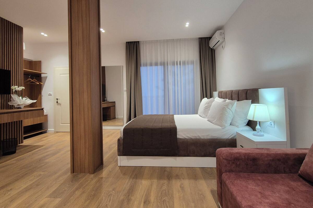 Una habitación de hotel moderna con una cama grande, una mesita de noche con lámpara, un sillón rojo, un armario de madera y un espejo.