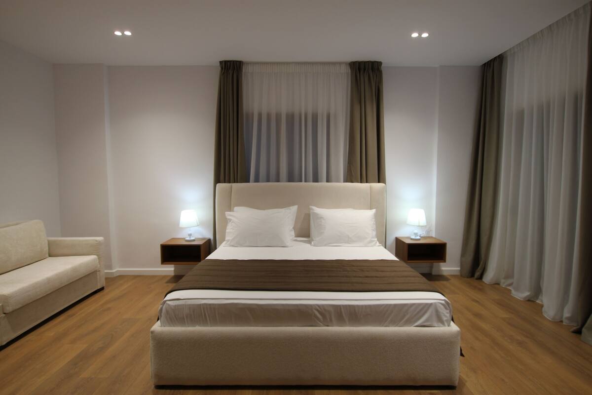 Una habitación de hotel moderna con una cama grande, una mesita de noche con lámpara, un sofá y un ventanal con cortinas.