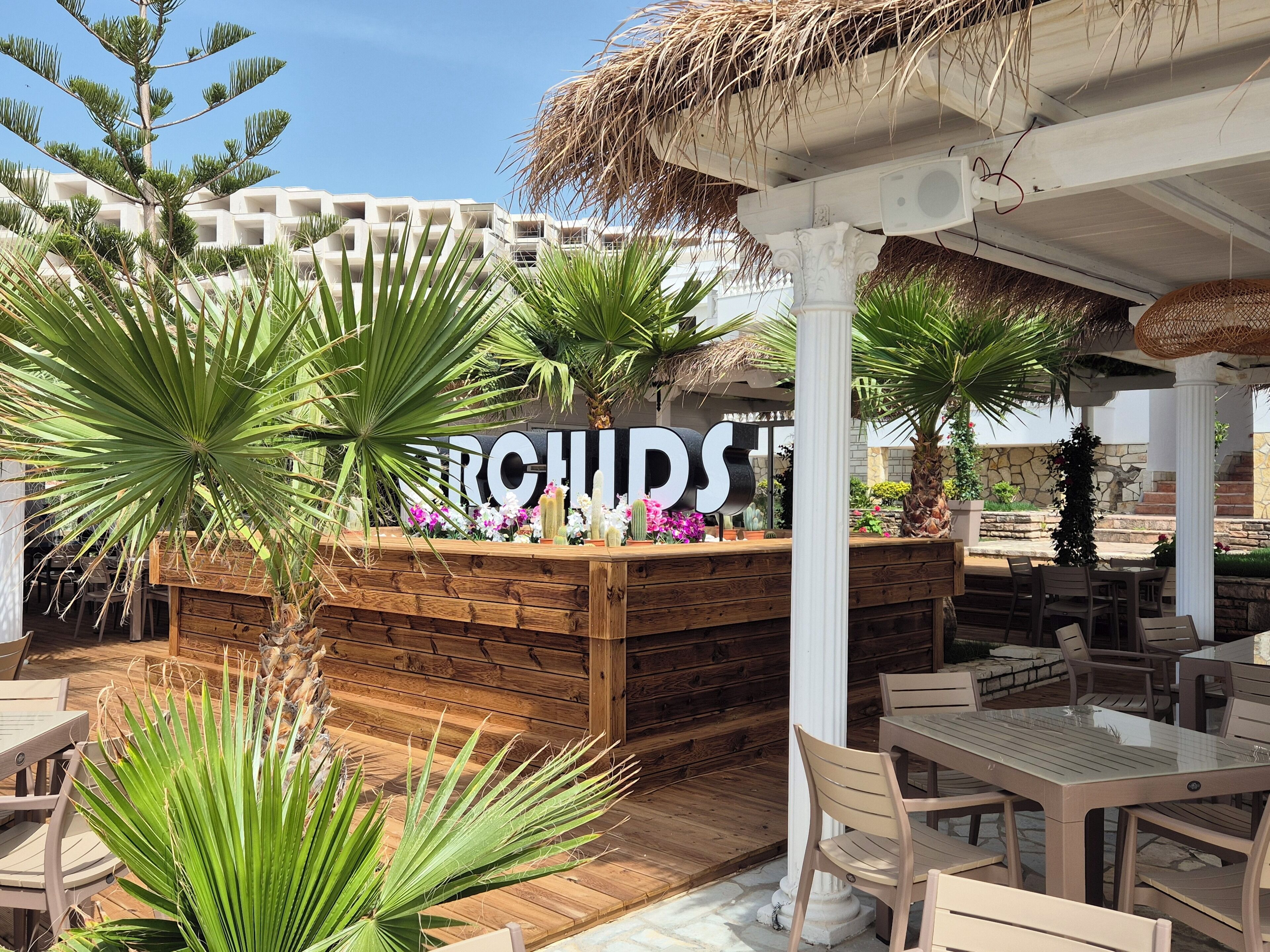 A wooden outdoor seating area with a thatched roof, surrounded by potted plants and a sign reading "CHURCHES".