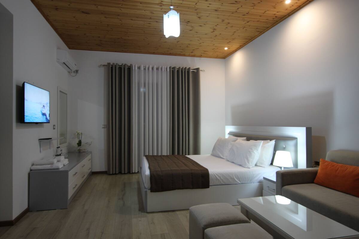 Una habitación de hotel moderna con techo de madera, una cama con sábanas blancas, un sofá gris, una mesita pequeña y un televisor en la pared.