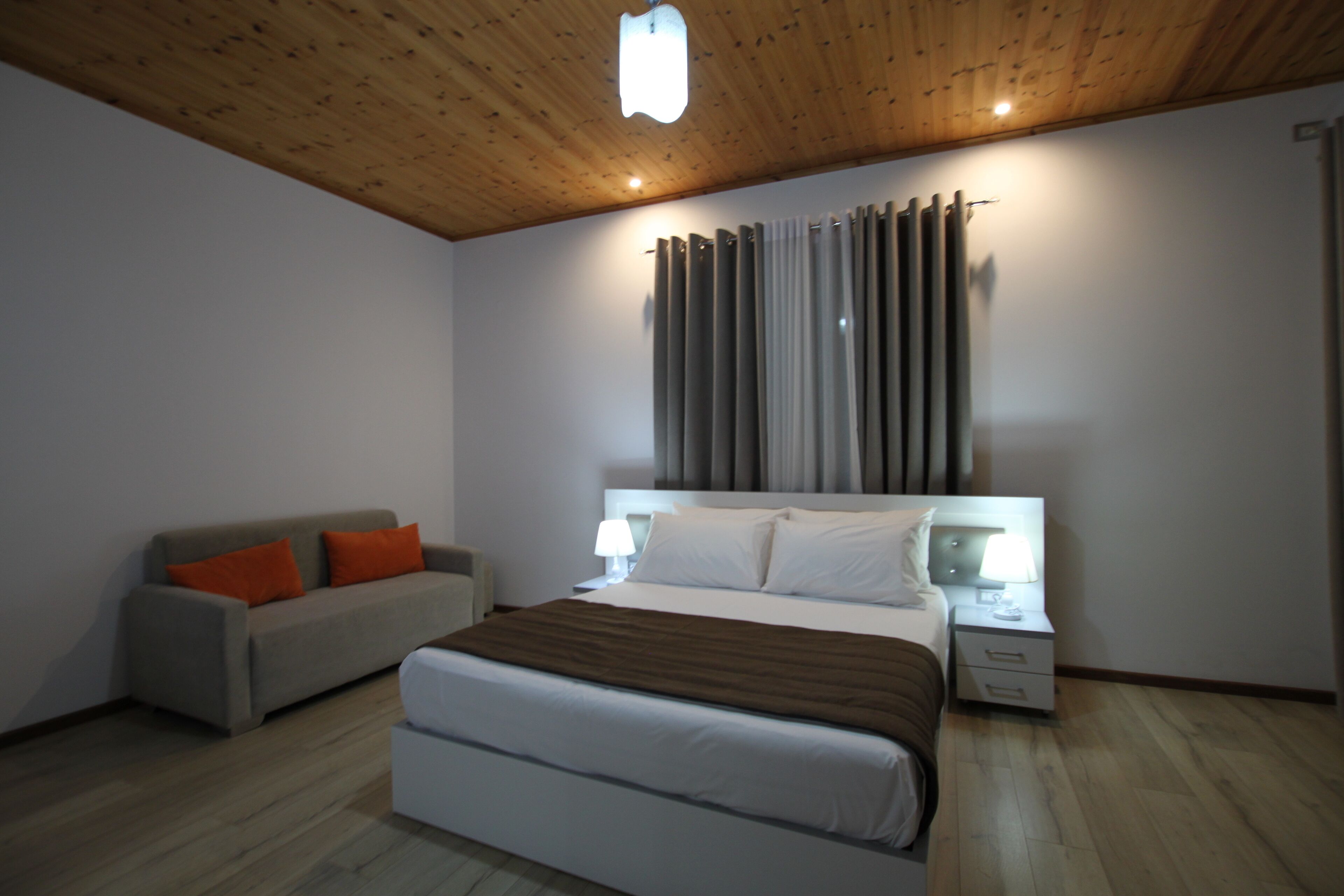 A modern hotel room with a wooden ceiling, a large bed with white bedding, a grey sofa with orange cushions, and a nightstand with a lamp.