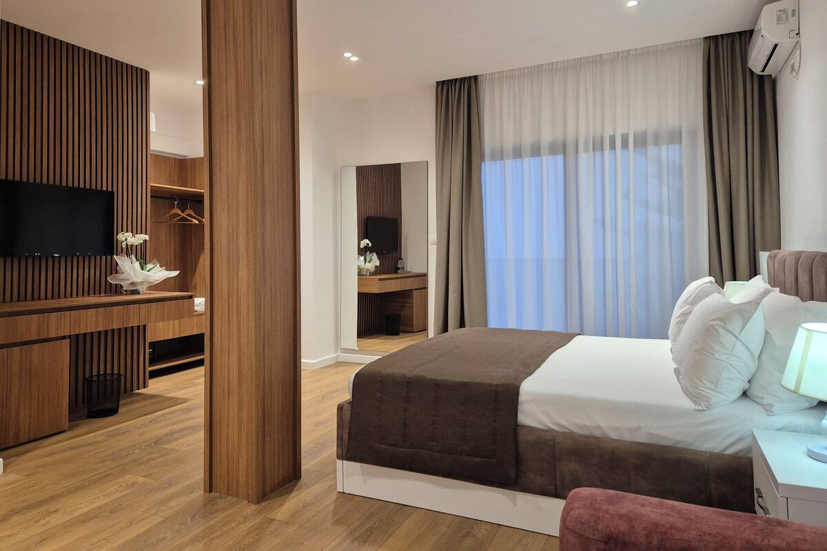 Una habitación de hotel moderna con pared de paneles de madera, televisión de pantalla plana, cama con cabecera marrón, mesita de noche con lámpara y un ventanal con cortinas.