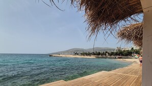 Une terrasse en bois qui mène à une plage aux eaux cristallines, avec une montagne lointaine et une vue sur la ville en arrière-plan.