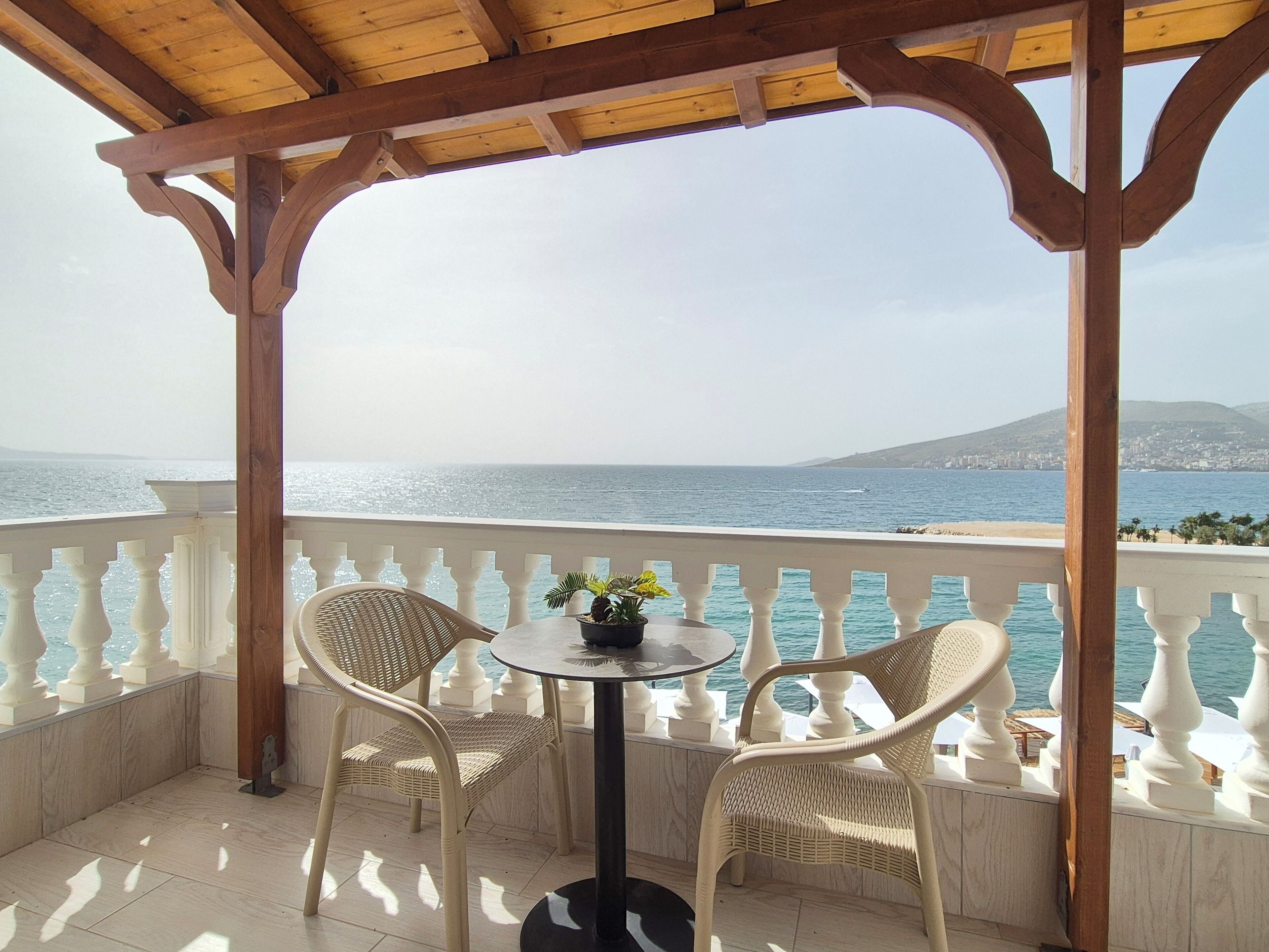 A covered outdoor seating area with a table and two wicker chairs, overlooking a coastal view.