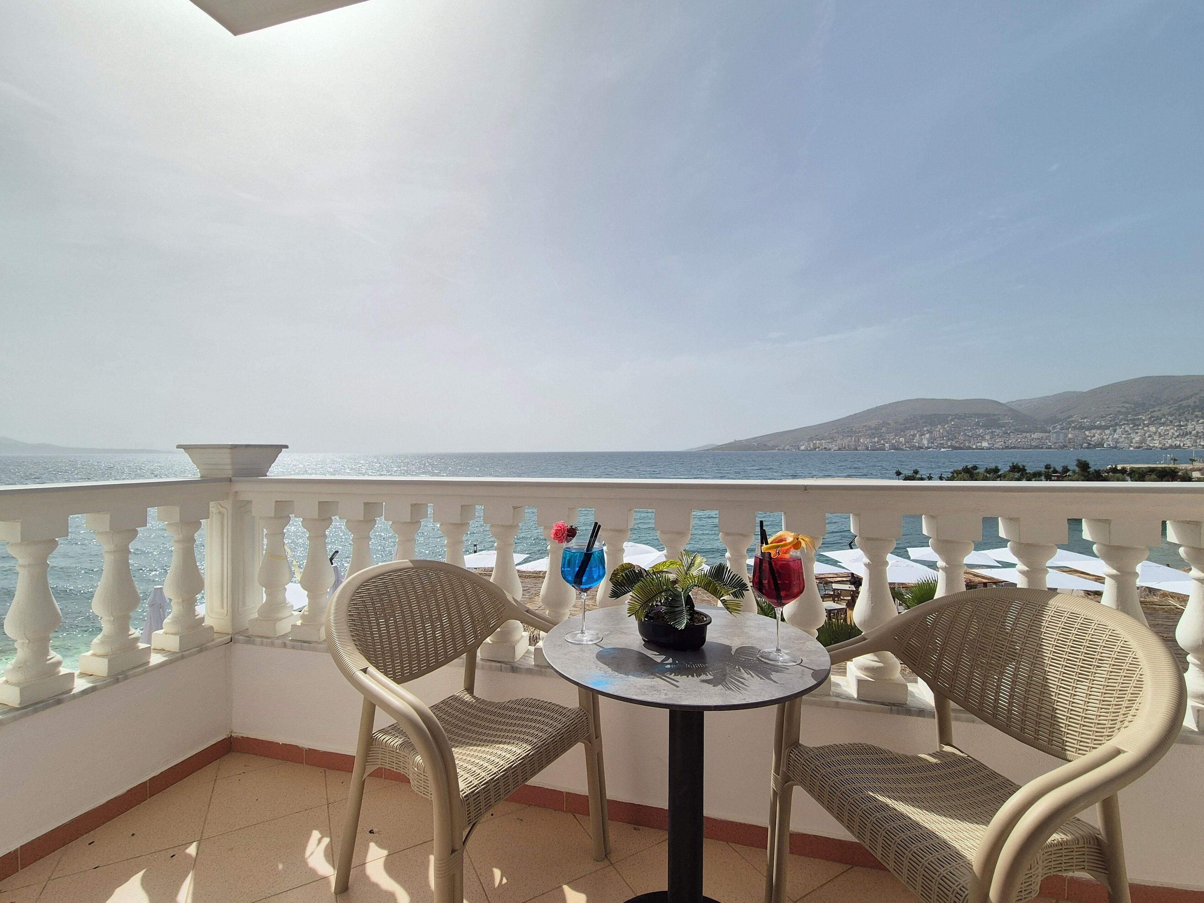 Un balcon avec deux fauteuils en osier et une petite table ronde, offrant une vue sur le paysage urbain de la côte.