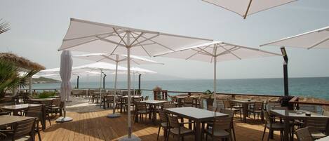 Un área de comedor al aire libre con mesas y sombrillas, con vista al mar.