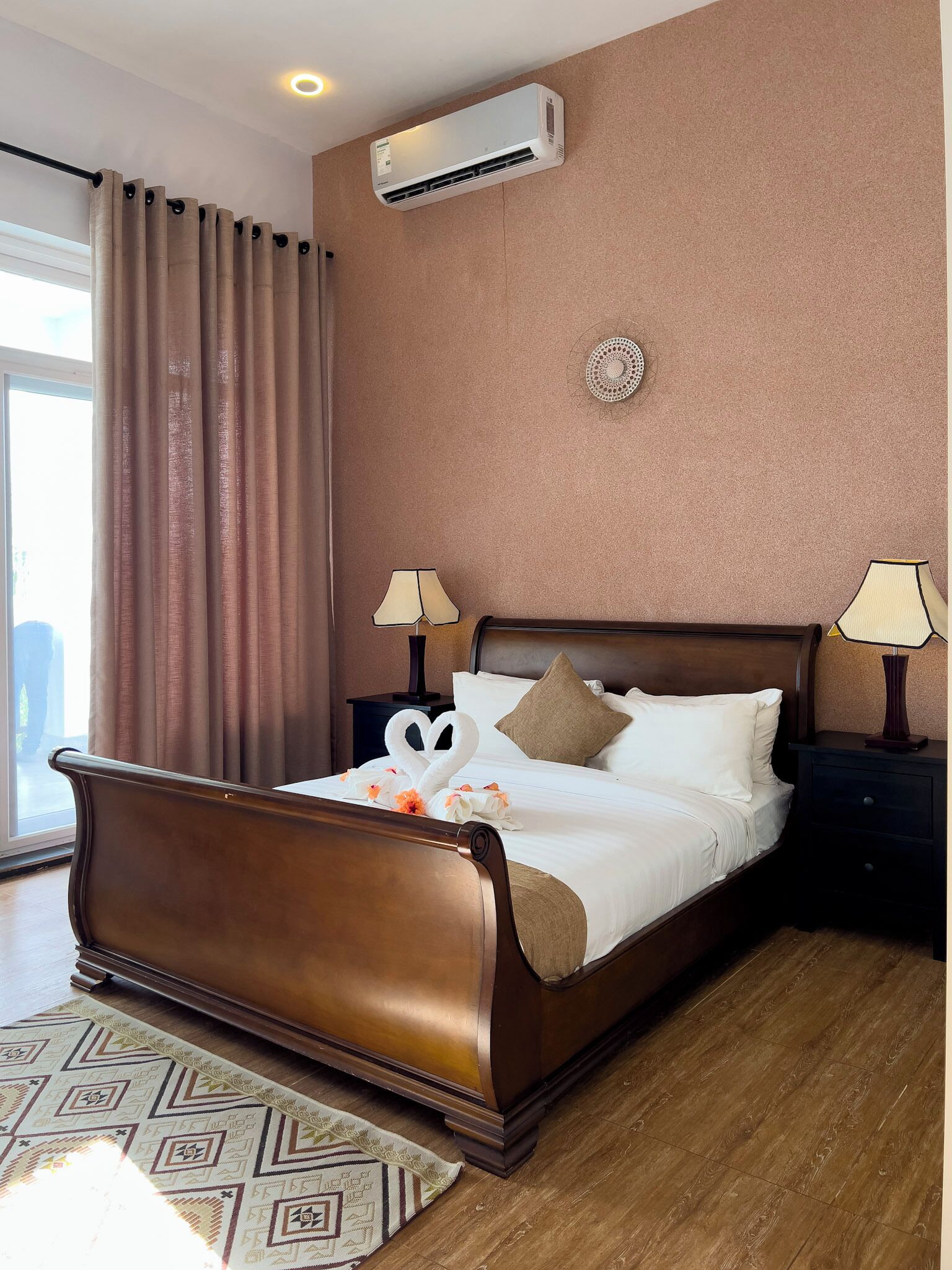 A hotel room with a wooden bed, two bedside lamps, a wall-mounted air conditioner, and a large window with curtains.