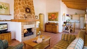 A cozy living room with a fireplace, wooden furniture, and a kitchen in the background. - SON MARGE (Campos)