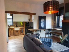 A modern living room with a grey sofa, a blue ottoman, and a dining area with a wooden table and chairs. A large painting is on the wall, and a hanging light fixture is above the dining table.