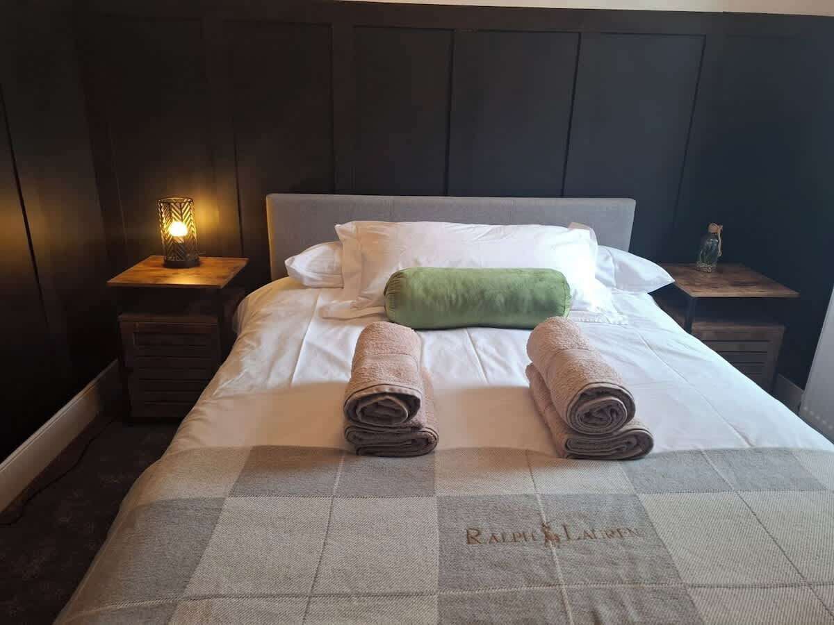 Een net opgemaakt bed met opgerolde handdoeken, een groene kussen en een geruit plaid.