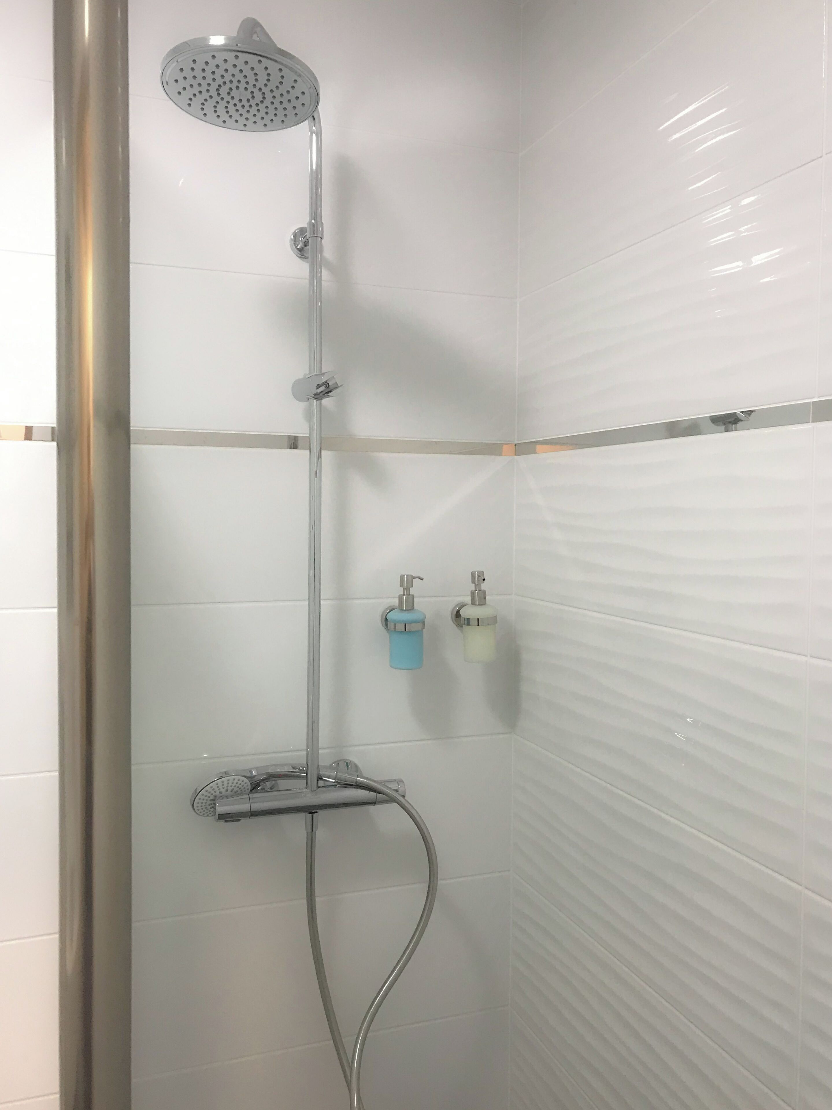 A shower with a handheld showerhead, a wall-mounted faucet, and a handheld shower hose. There are two soap dispensers on the wall.