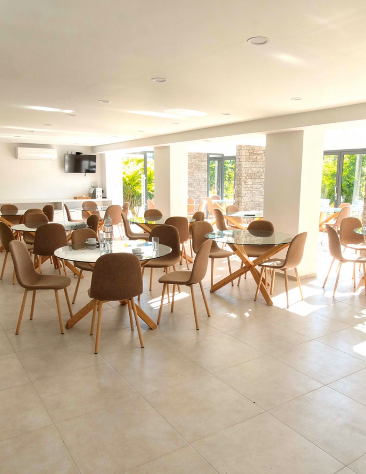 A spacious dining area with multiple round tables and chairs, a flat-screen TV mounted on the wall, and large windows allowing natural light.