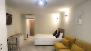 Una habitación compacta con una cama, un sofá amarillo, un escritorio con silla y un televisor en la pared.