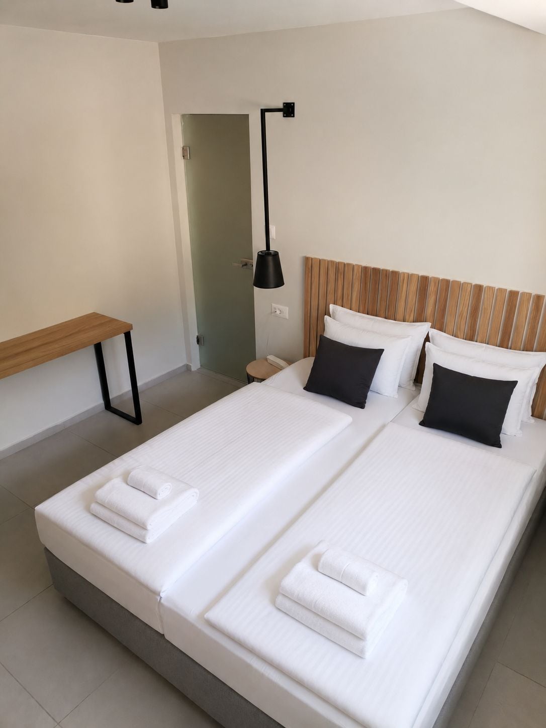 A double bed with white linens and black pillows, a bedside table, a lamp, and a wooden headboard.
