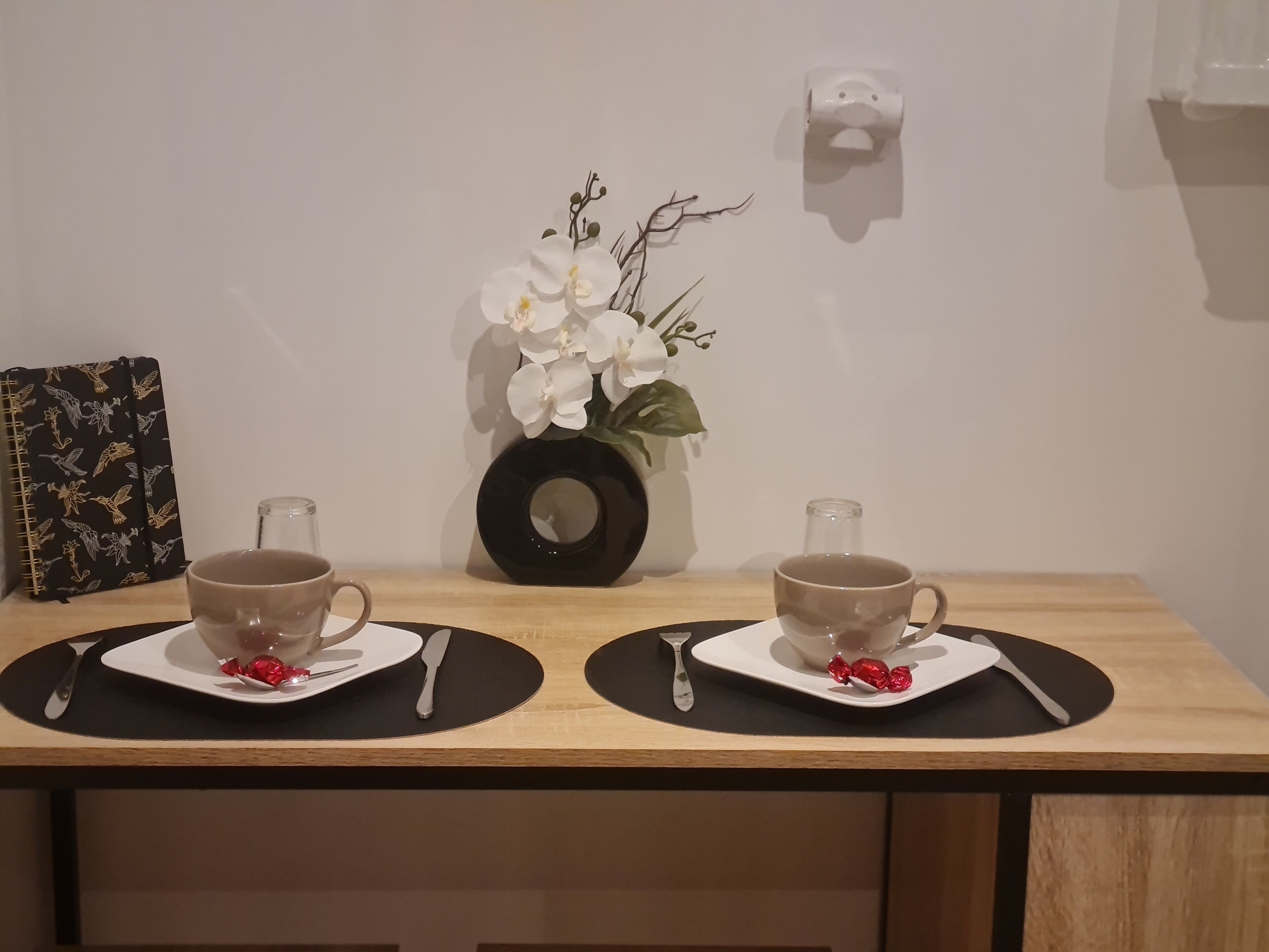 Une table à manger avec deux tasses de café, un vase de fleurs blanches et une horloge décorative au mur.