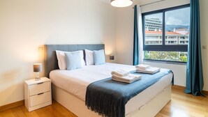 Quarto de hotel moderno com cama grande, abajures, vista de edifícios pela janela e roupa de cama bem arrumada com toalhas.