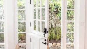 Una habitación con una puerta blanca que da a un jardín, una silla de mimbre y suelo de baldosas.
