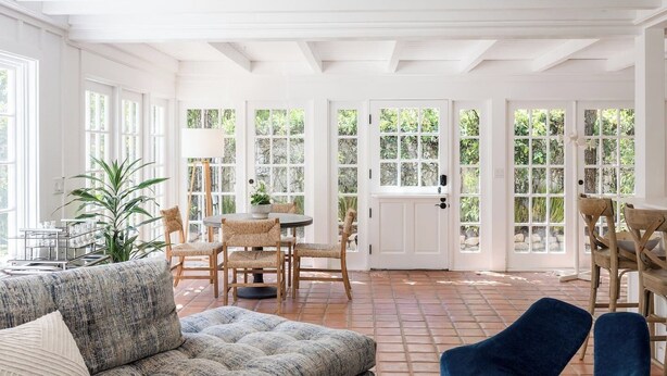 Una habitación luminosa e inundada de sol, con ventanas amplias, una mesa redonda con sillas, un sofá y una planta en maceta.