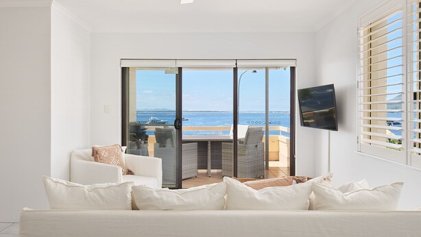 A modern living room with a white sofa, a flat-screen TV mounted on the wall, and a view of the sea through a sliding glass door. - Del Mar - 1 bed option (Shoal Bay)