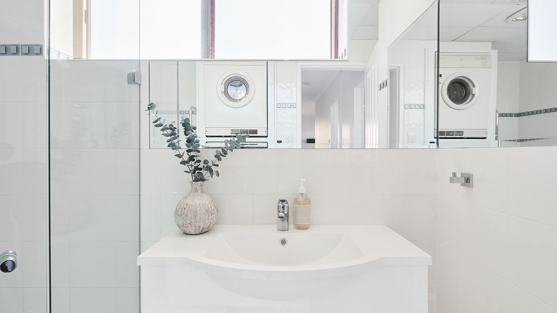A modern bathroom with a white sink, a mirror reflecting a washing machine, and a potted plant.