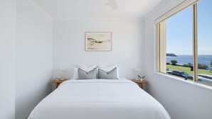 A bedroom with a large bed, white walls, a framed artwork above the bed, and a view of the sea through a large window. - Del Mar - 1 bed option (Shoal Bay)