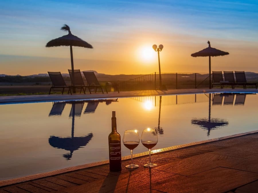 Una piscina infinita con sillas de descanso y sombrillas, una botella de vino y dos copas al borde.